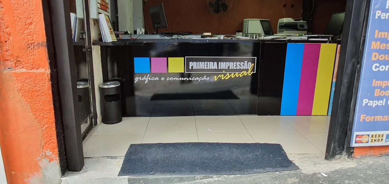 Balcão da Primeira Impressão: painel preto com o nome da gráfica e faixas ciano, magenta e amarelo, impressoras ao fundo.